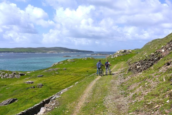 two people walking together with bikes in green field viewing mountain and blue sea under white and blue sky during daytime